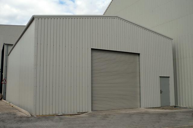 A large grey building with a roller door and personal access door at the front beside an even larger grey building